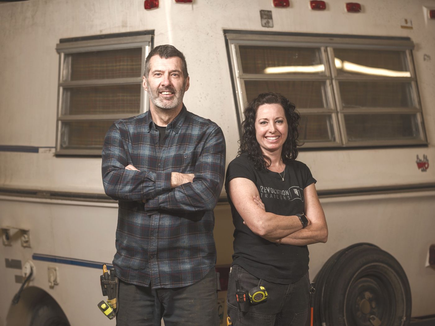 Stu and Megan in front of a vintage trailer inside their shop.